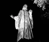 The image shows a black and white photo of a statue of a robed figure with outstretched arms illuminated from below against a dark background