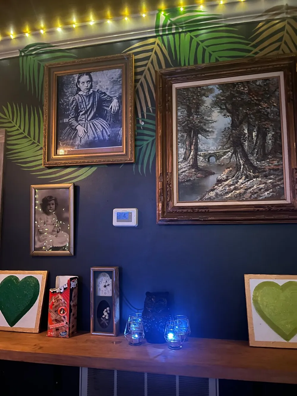 The image displays a stylishly decorated wall with an eclectic mix of artwork and objects including framed pictures and paintings illuminated by string lights above a wooden shelf adorned with unique items and glowing bulb lights
