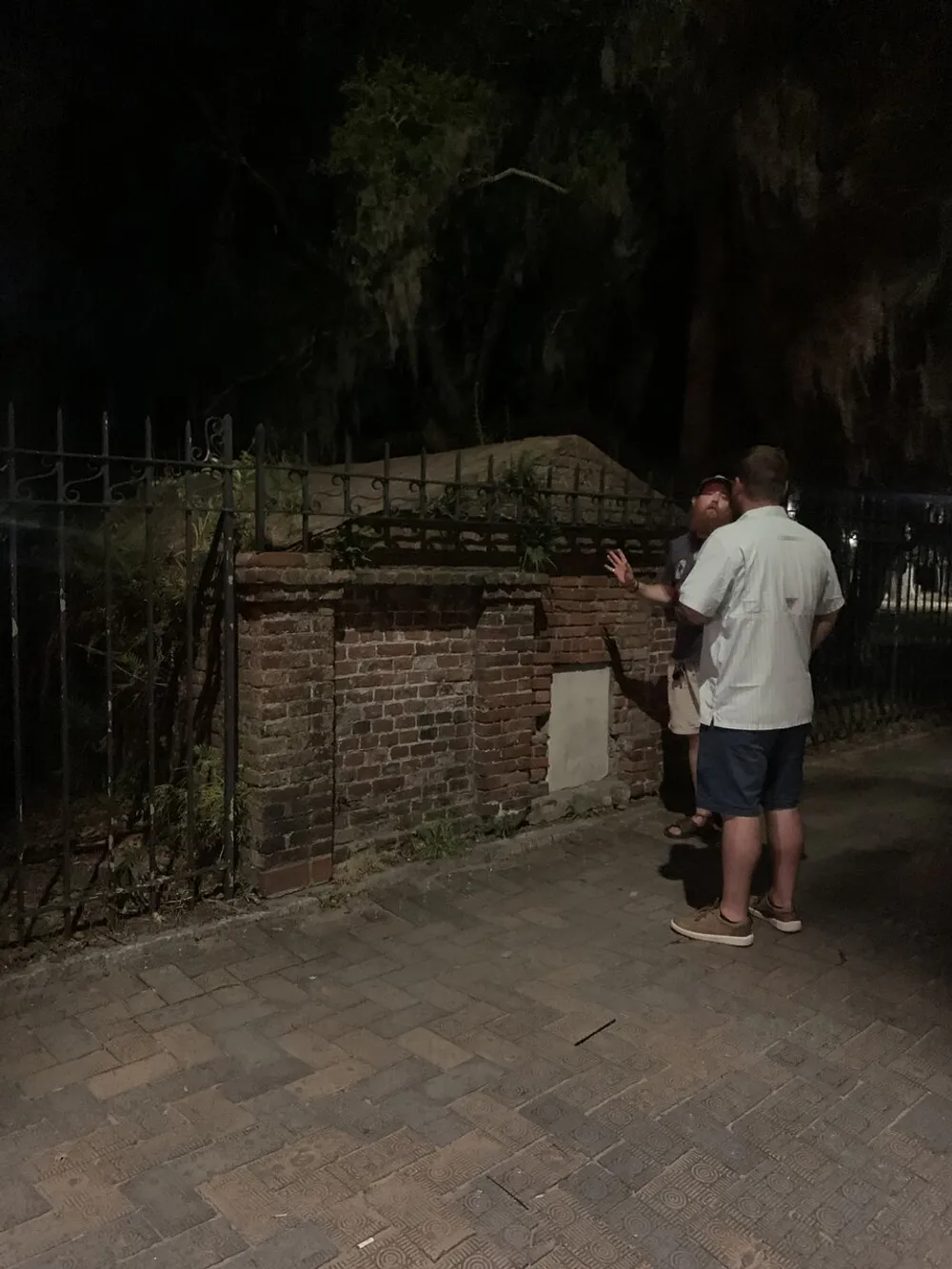 Two individuals are standing at night by a brick structure with overhanging Spanish moss seemingly in a discussion or examination of the place