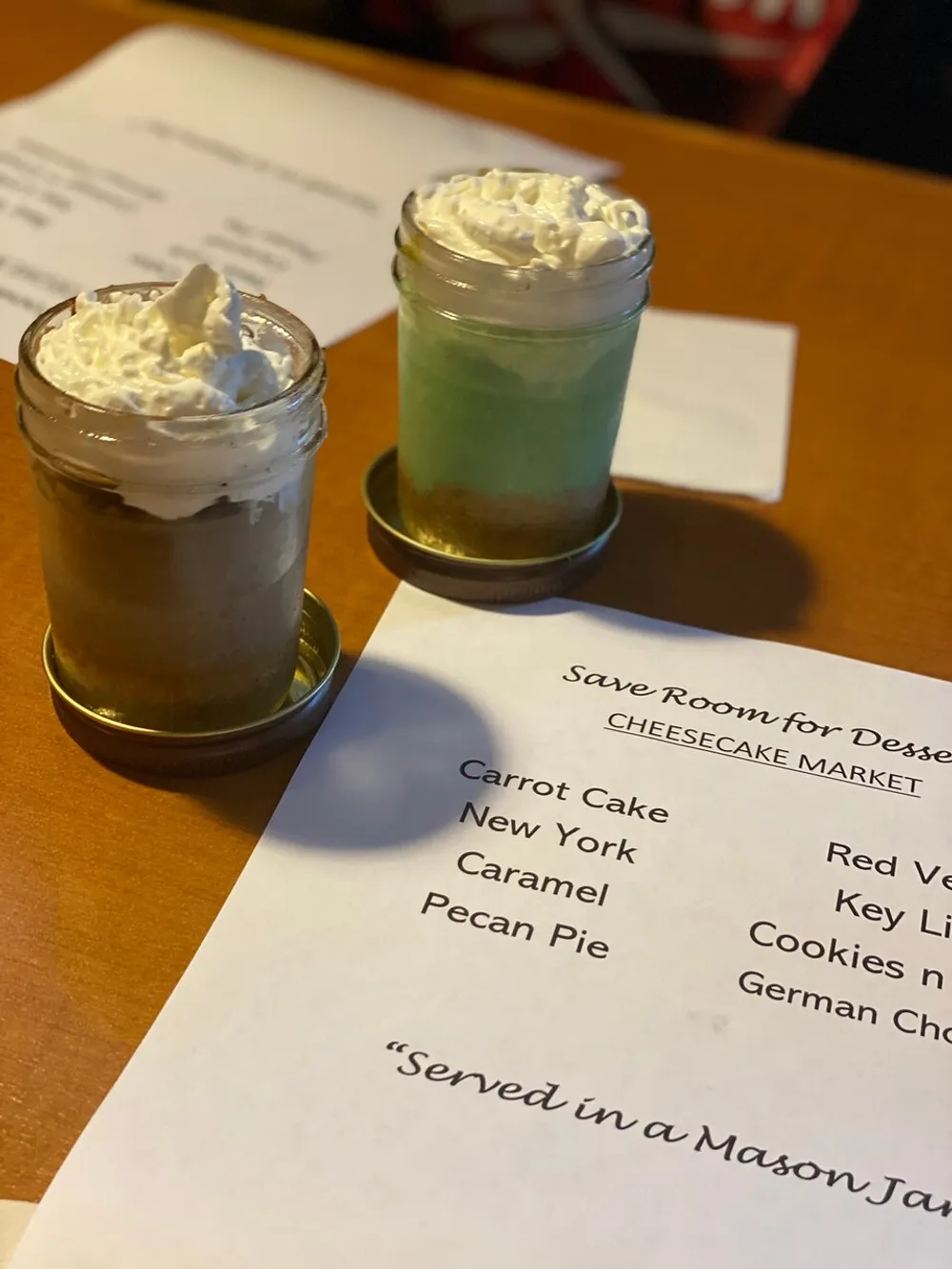 The image displays two mason jars filled with layered desserts topped with whipped cream situated in front of a dessert menu which indicates they are part of a Cheesecake Market selection