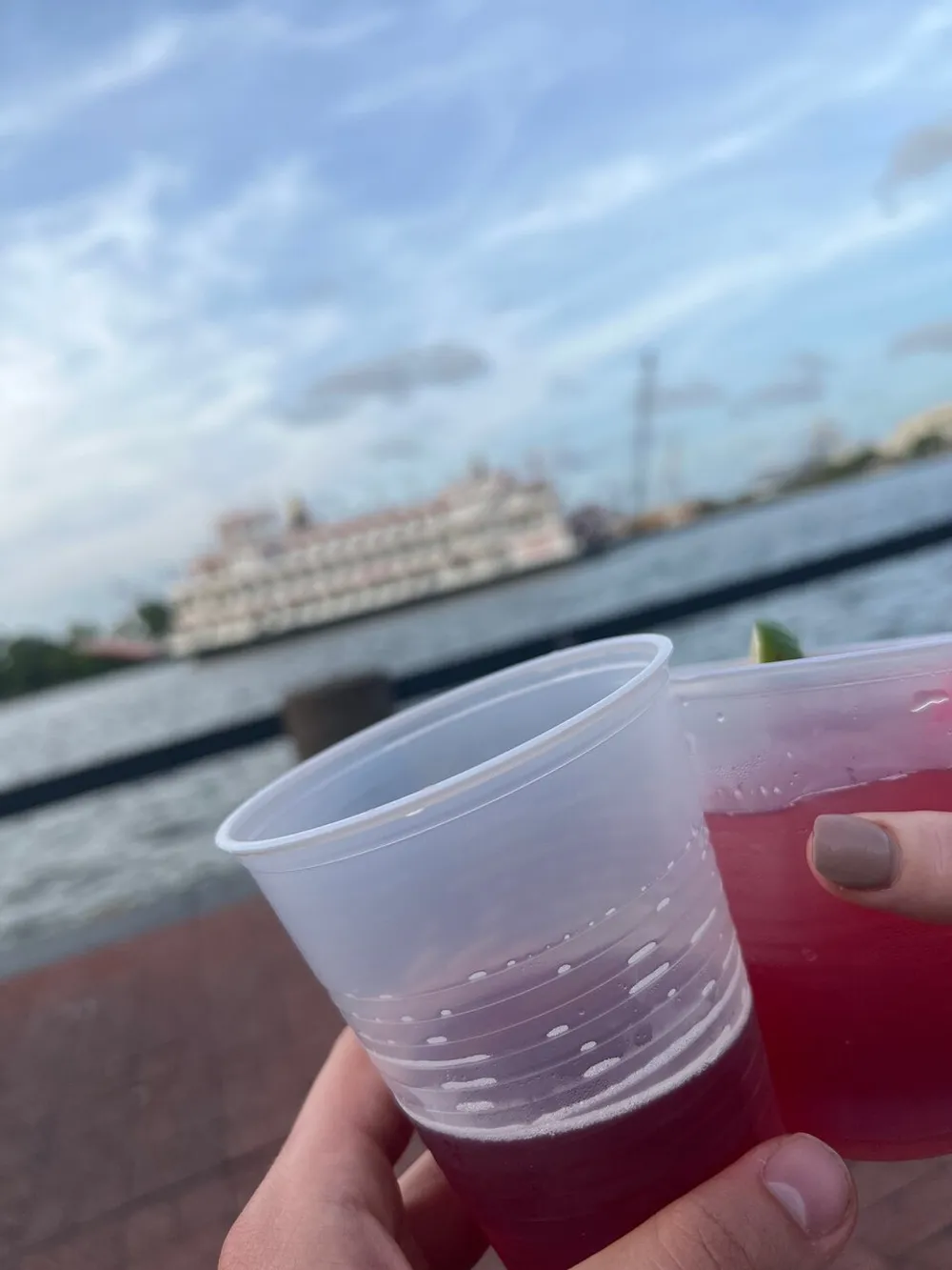 A person is holding two plastic cups of beverages with a blurred view of a waterway and a boat in the background