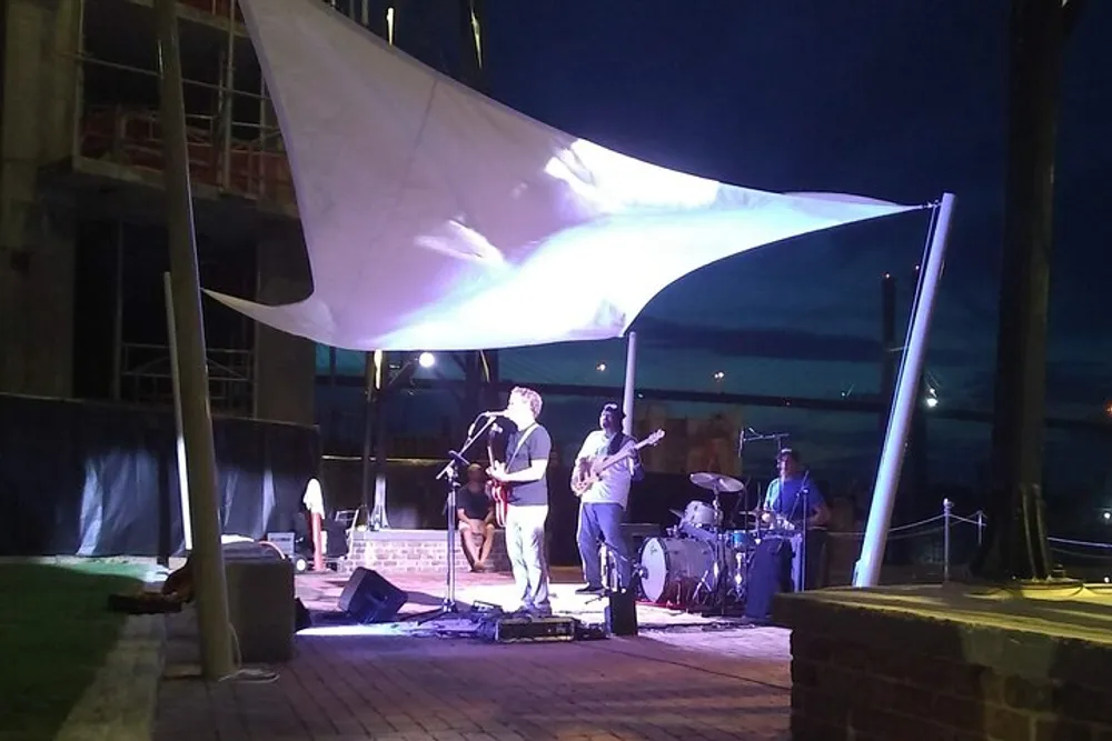 A band performs on an outdoor stage at night under a large white canopy with building construction visible in the background