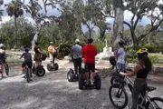 A group of people on a mixture of segways and bicycles gathered in a park with Spanish moss-draped trees, listening to someone who appears to be a guide.