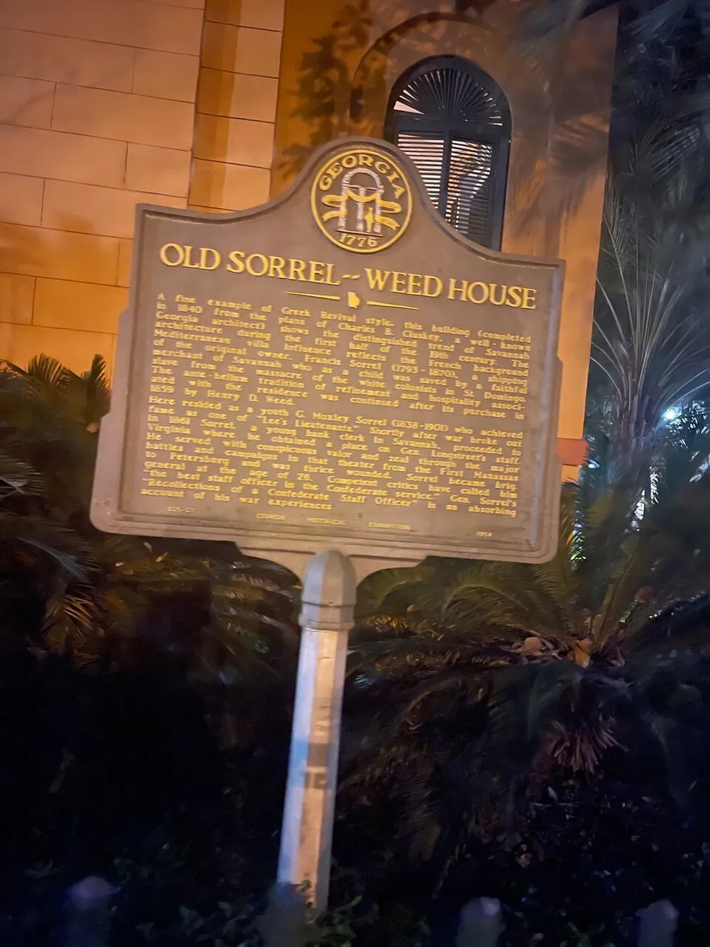 The image shows a historical marker for the Old Sorrel-Weed House a Greek Revival-style building located in Savannah Georgia illuminated by a warm light at night and surrounded by foliage