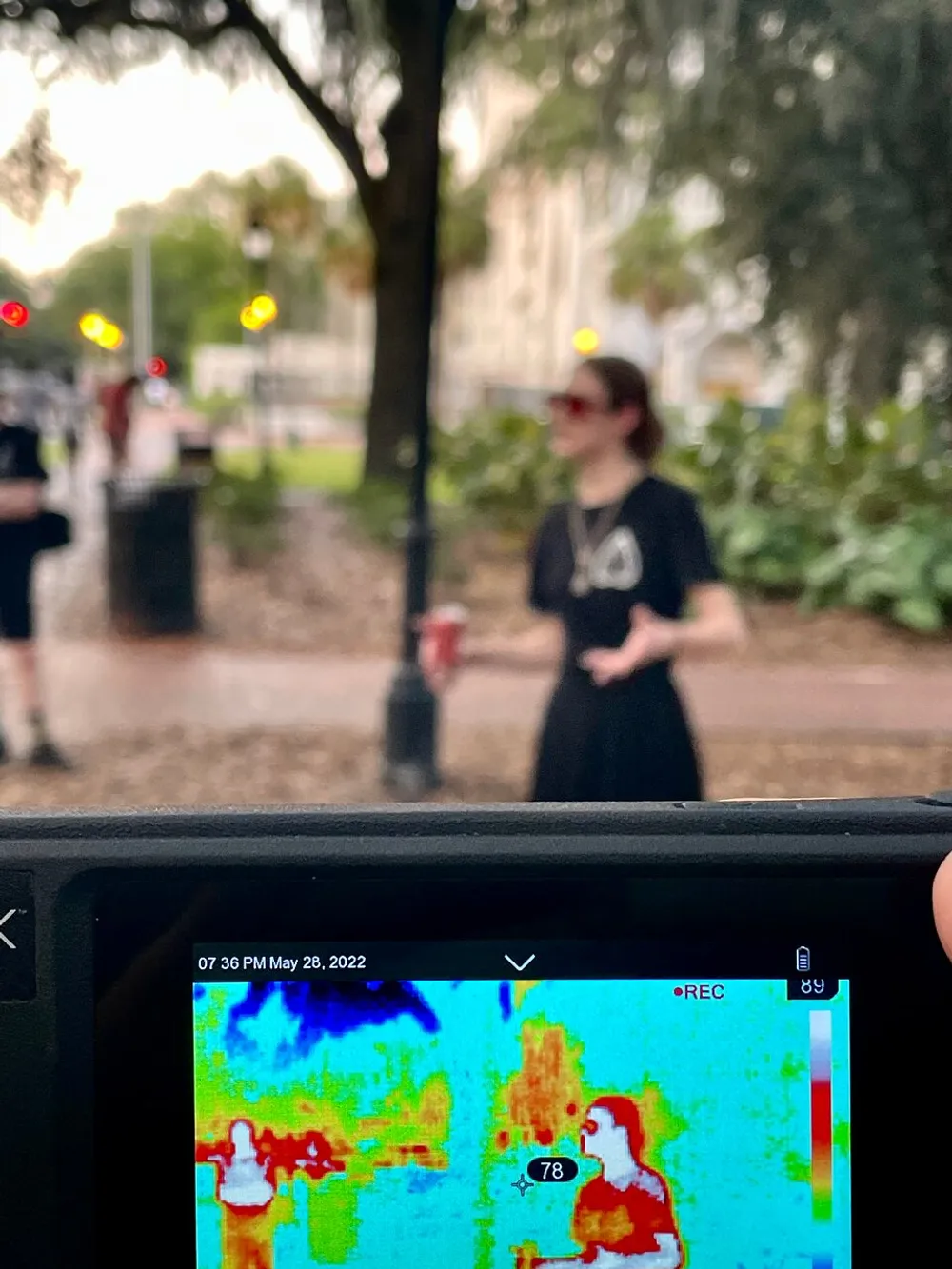 The image shows a person holding a camera with a thermal viewfinder that is focused on another person standing in the park with a time stamp indicating the photo was taken on May 28 2022 at 736 PM