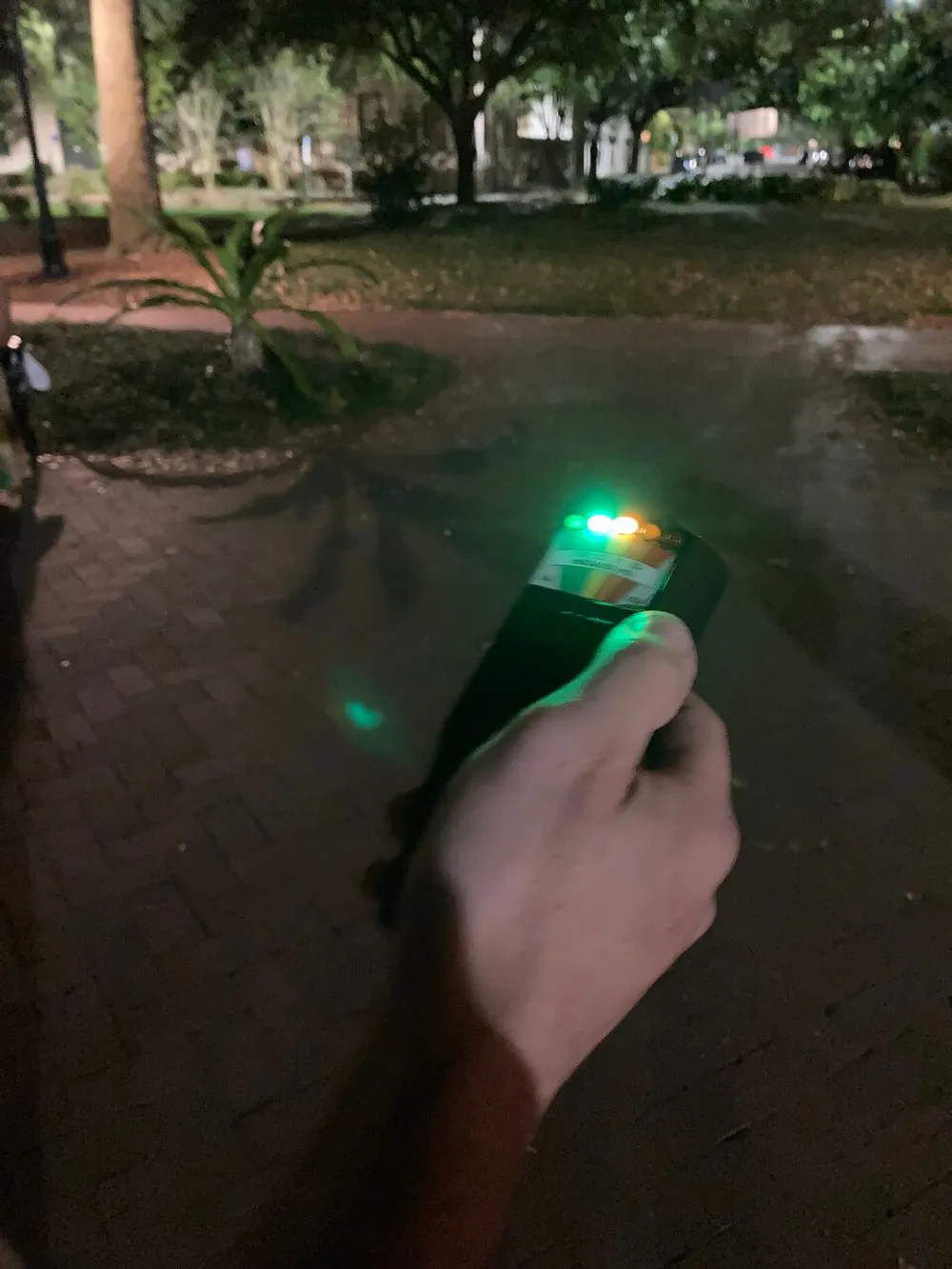 A person is holding a device with glowing lights possibly a measurement tool or electronic gadget in a dimly lit outdoor setting at night