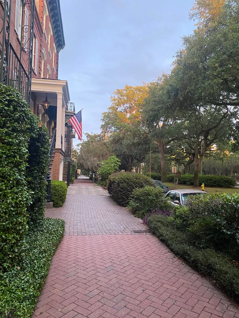 The image shows a tranquil brick-paved street lined with well-maintained hedges and trees alongside elegant buildings with an American flag hanging from a balcony suggesting a peaceful residential area