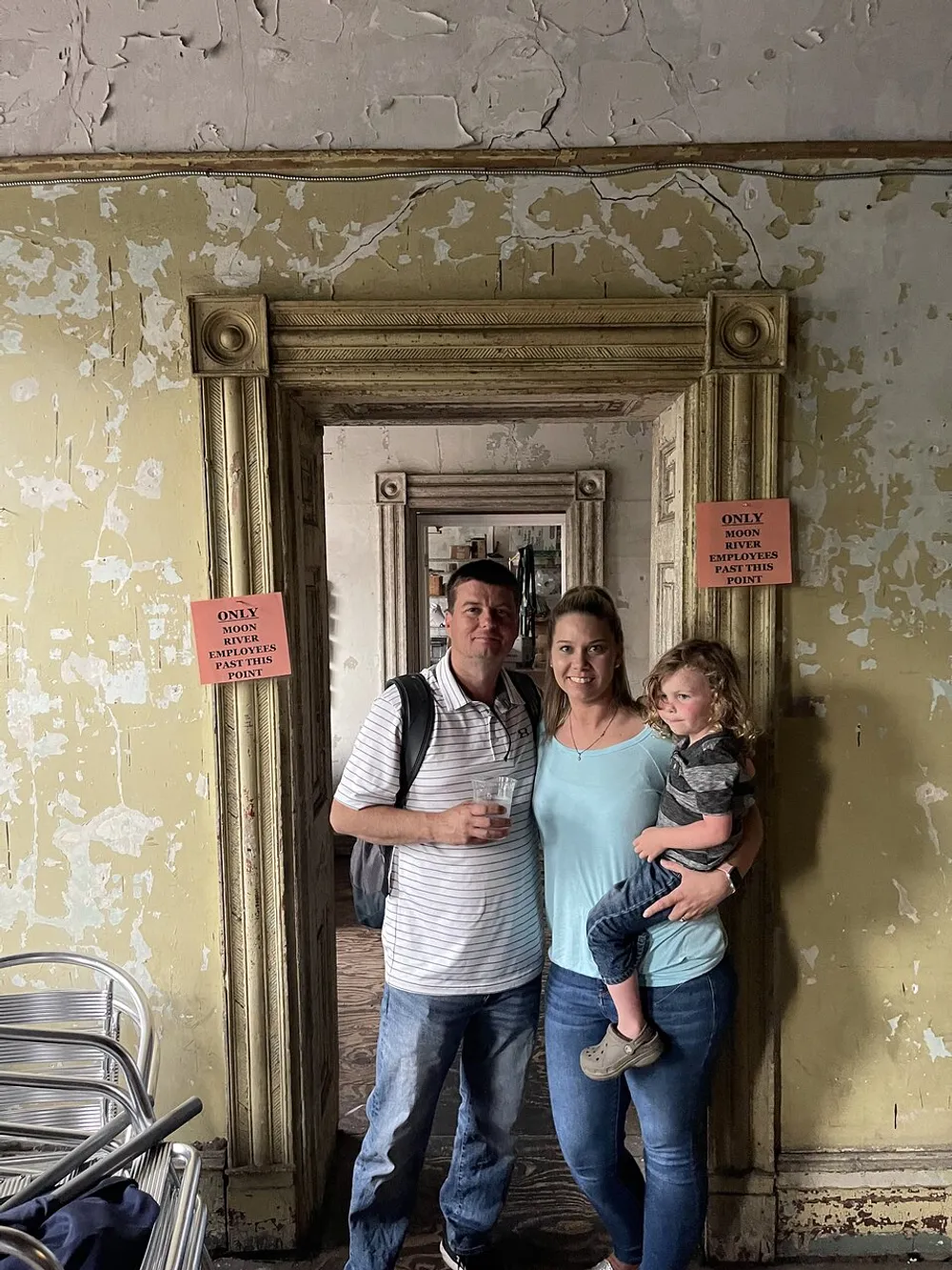 Three people are standing together smiling in front of a peeling wall inside a room with an ornate mirror and signs that say ONLY EMPLOYEES PAST THIS POINT