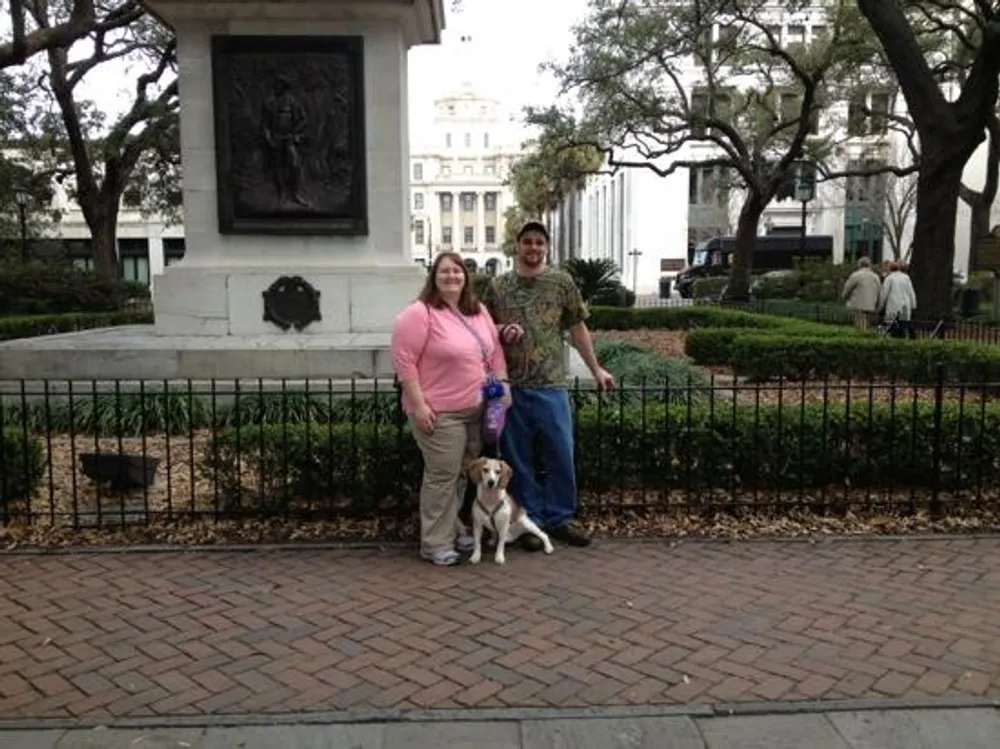 A man and a woman pose for a photo with their dog in what appears to be a park with a statue and a building in the background