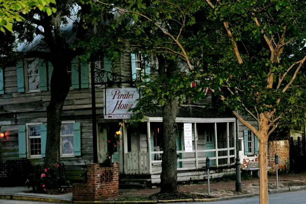 The image shows a rustic weathered building with a sign reading Pirates House surrounded by trees in what appears to be the soft light of either dawn or dusk