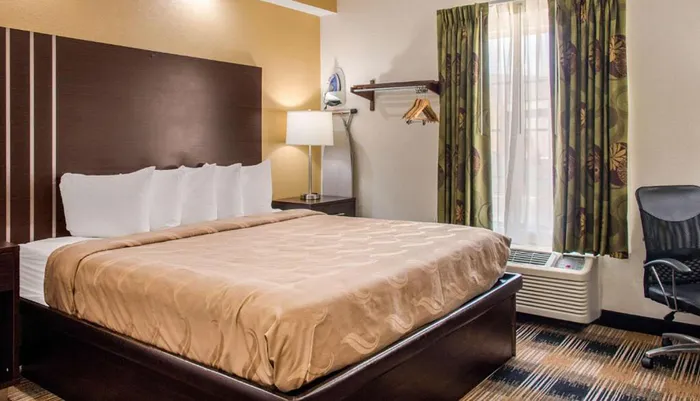 The image shows a neatly made bed with white pillows in a hotel room that also contains amenities like a side table with a lamp an ironing board and a window with curtains