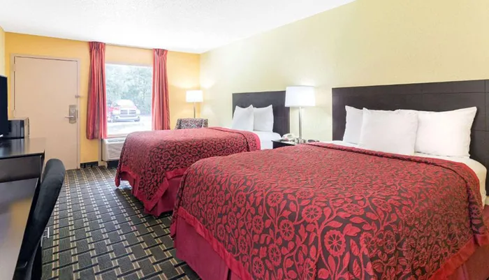 The image shows a standard hotel room with two double beds covered in red patterned bedspreads simple furnishings a flat-screen TV and a window with a view of a vehicle outside