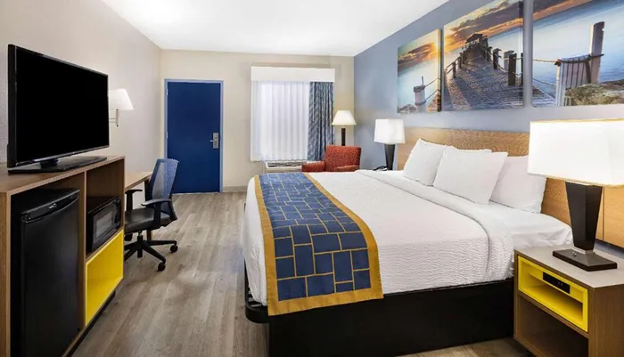 The image shows a modern hotel room with a large bed a desk with a chair a flat-screen TV and a decorative wall picture above the headboard