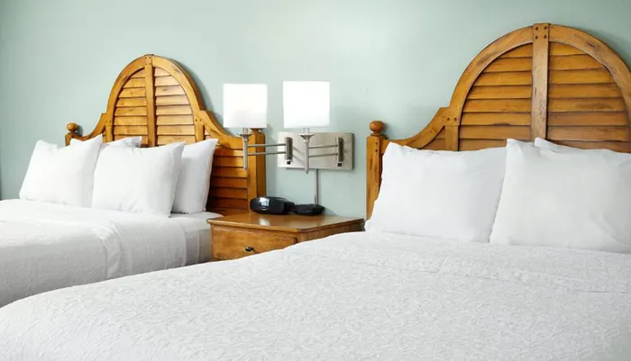 This image shows a neatly made bedroom with two twin beds featuring unique curved wooden headboards white bedding and a shared nightstand with a lamp and a clock