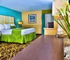 The image displays a bright and colorfully decorated hotel room featuring a bed with a green bedspread complemented by aqua walls patterned carpet and a vase with tall greenery on a wood-toned surface in the foreground