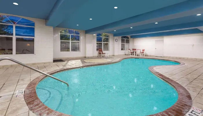 The image shows an inviting indoor swimming pool with a kidney-shaped design large windows and a sitting area all under a blue ceiling