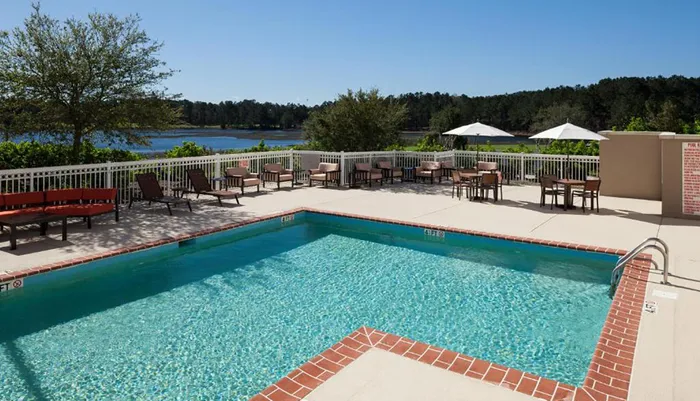 The image shows an outdoor swimming pool with deck chairs and tables on the surrounding patio overlooking a serene lake under a clear sky