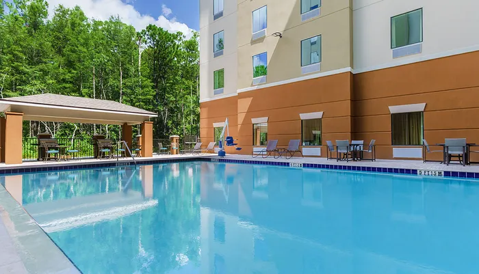 The image shows an outdoor hotel swimming pool with a clear blue water surrounded by lounge chairs tables with seating areas under a gazebo and bordered by a forested area all adjacent to a multi-storey hotel building