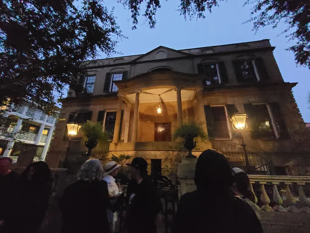 A group of people is gathered in the twilight outside a stately old house with illuminated windows and a lit porch lantern creating a mysterious atmosphere