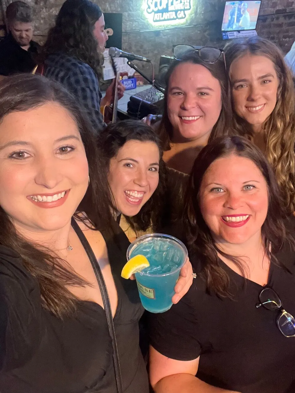 A group of smiling women is posing for a selfie at a lively bar with a musician performing in the background