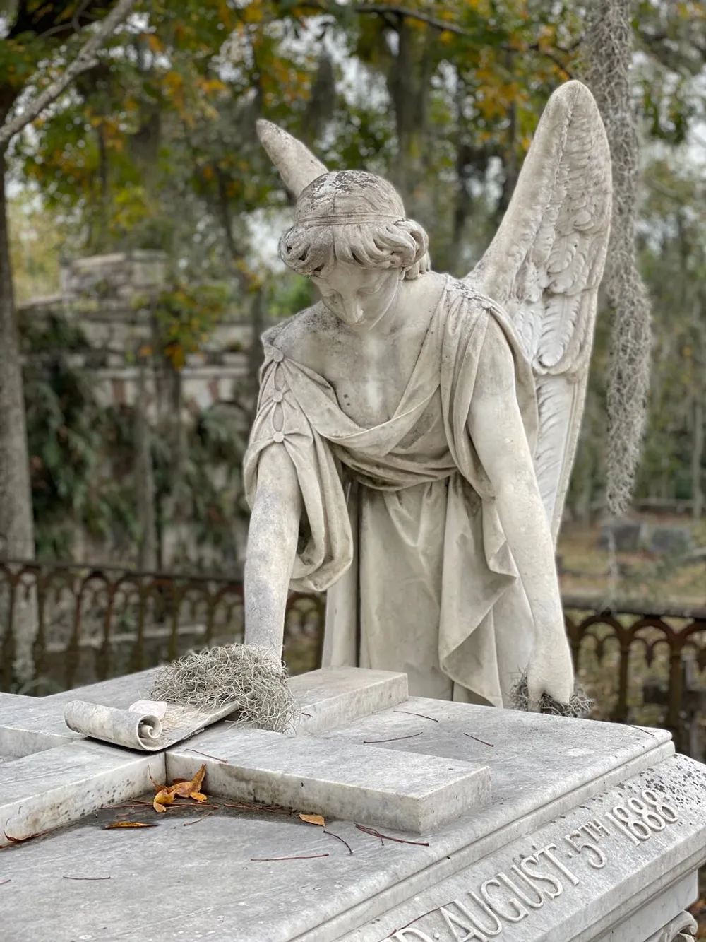 The image shows a weathered stone statue of an angel leaning sorrowfully over a tombstone with Spanish moss and Autumn leaves adding to the serene yet somber atmosphere of a cemetery