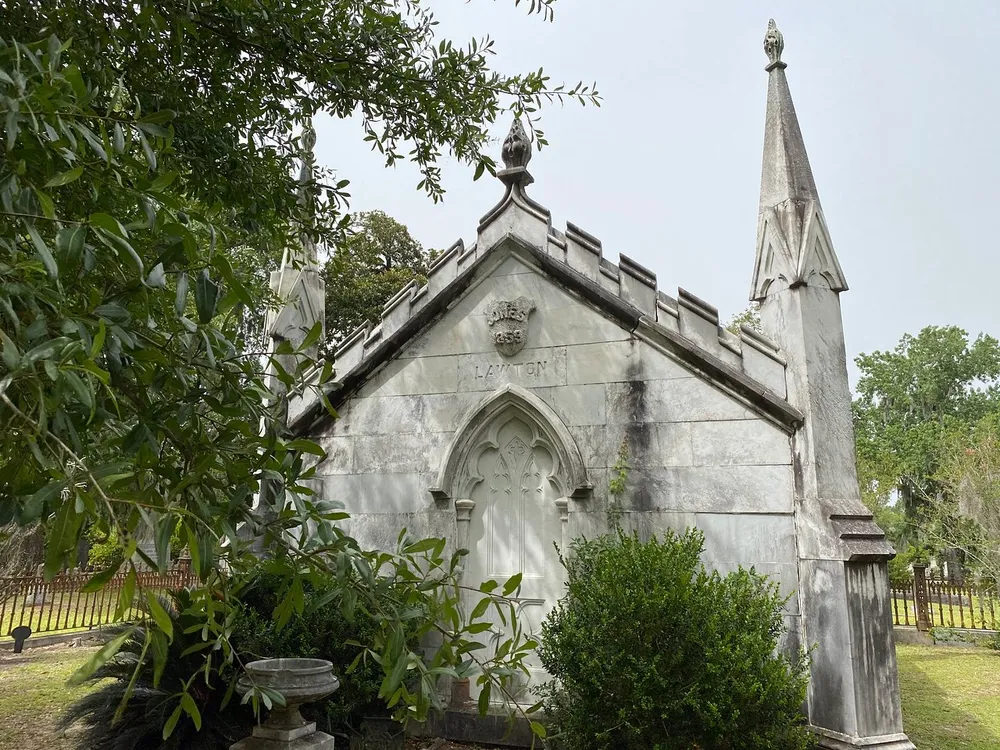 The image shows an ornate gothic-style mausoleum in a tranquil cemetery setting partially obscured by lush green foliage