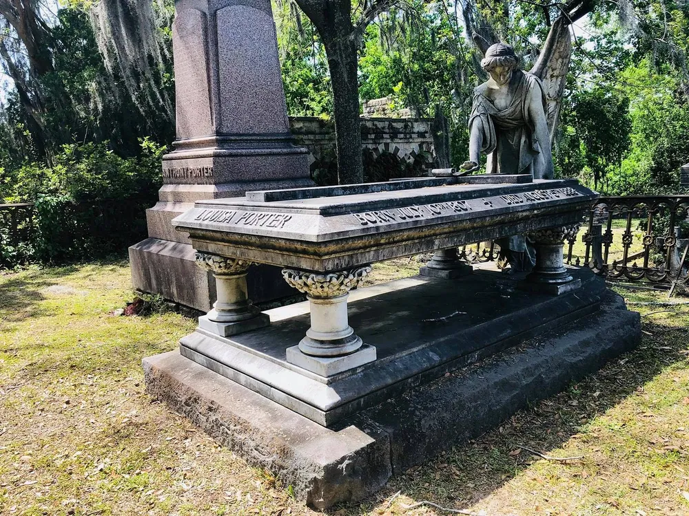 The image shows an intricate and ornate grave marker with a sculpture of a mourning angel leaning over a stone slab surrounded by nature and iron fencing