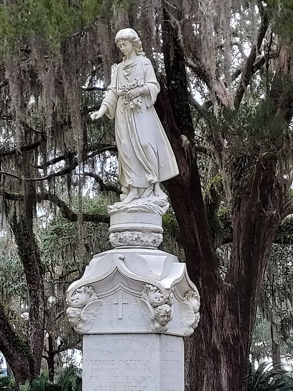 The image displays a detailed statue of a standing figure with outstretched hand positioned atop a memorial featuring cherubic faces and inscriptions all set against a backdrop of Spanish moss-draped trees