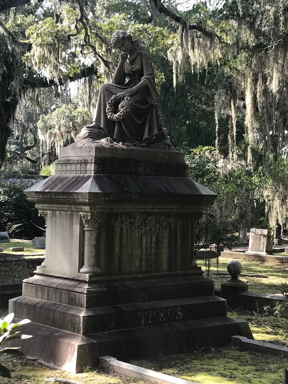 A solemn statue of a seated mourning figure holding a wreath atop a grave surrounded by Spanish moss-draped trees in a tranquil cemetery setting