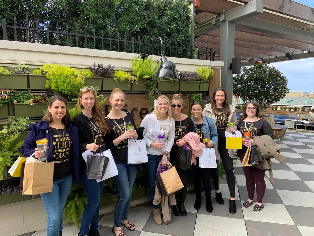 A group of happy individuals wearing matching t-shirts that say A Little Party Never Hurt Nobody are posing for a photo with various shopping bags and drinks in their hands standing in an outdoor setting with a vertical garden in the background