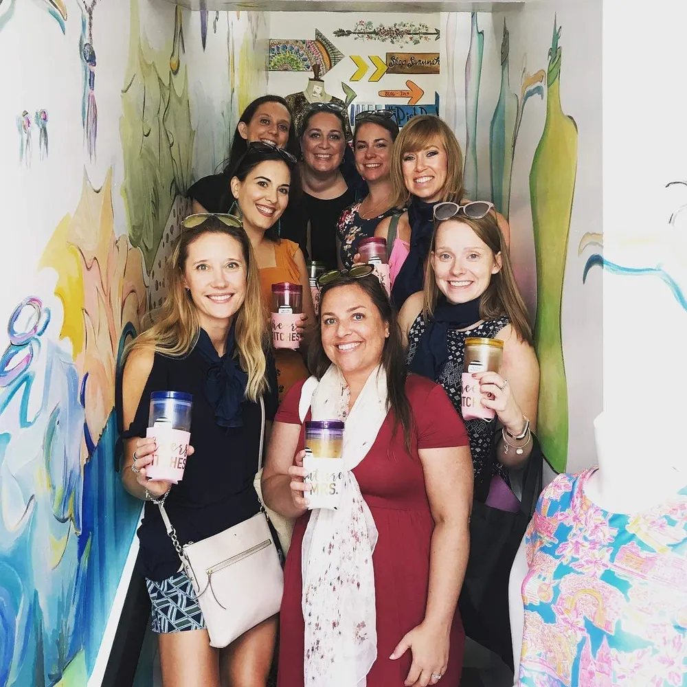 A group of smiling women is posing for a photo in a colorful room each holding a personalized drinking cup