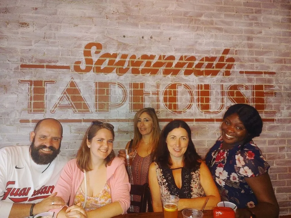 A group of five smiling people is posing for a photo at the Savannah Taphouse with a rustic brick wall backdrop accentuating the establishments name