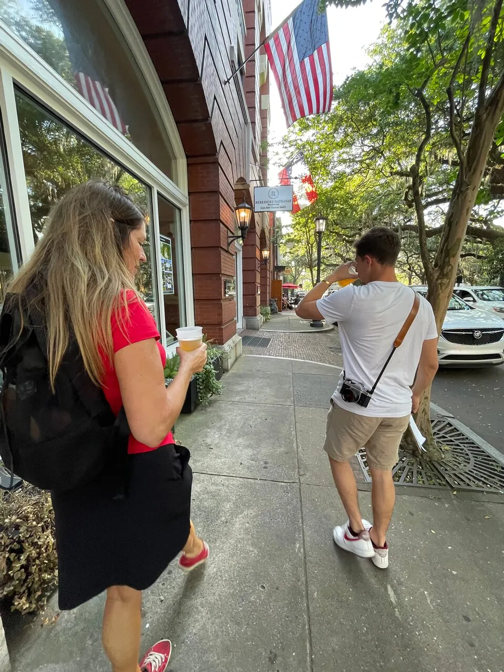 A man and a woman are walking on a sidewalk with the woman holding a coffee cup passing by a building that is flying American flags on a sunny day