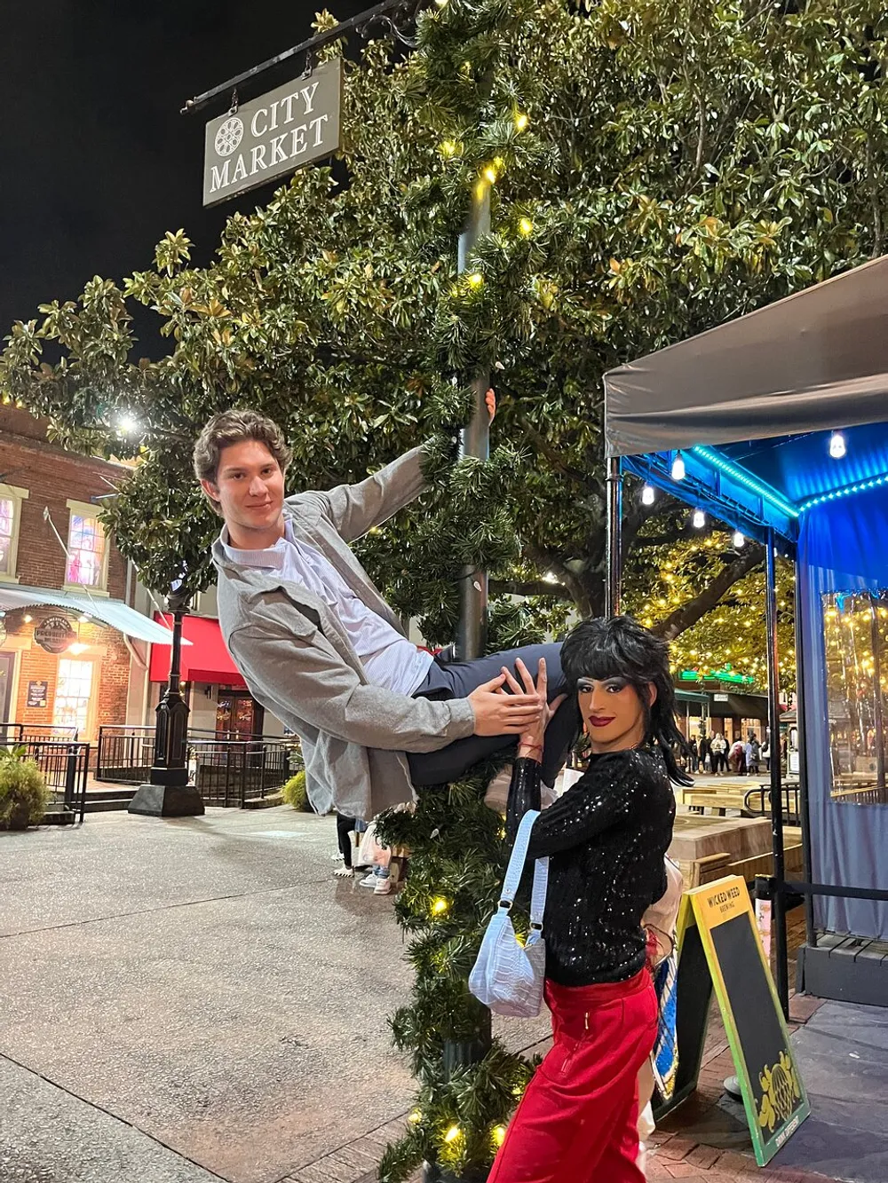 A person is playfully climbing a lamppost decorated with greenery while another person stands next to them posing with a smile on a city street at night