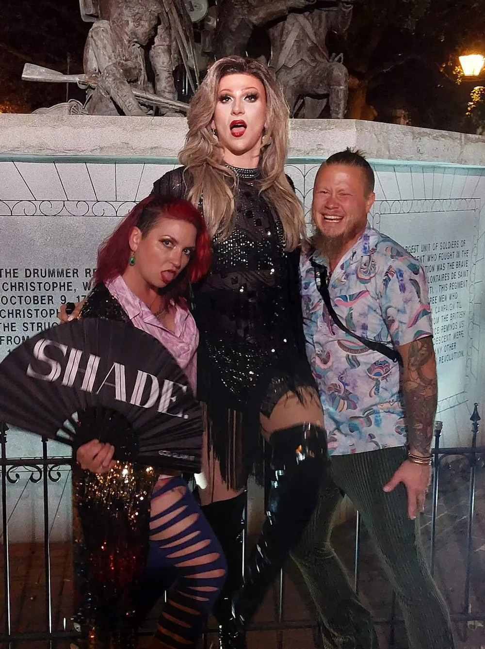 Three people are posing playfully at night with the central figure in elaborate drag attire and makeup while one holds a fan labeled SHADE and the other wears a patterned shirt in front of a sculpted memorial