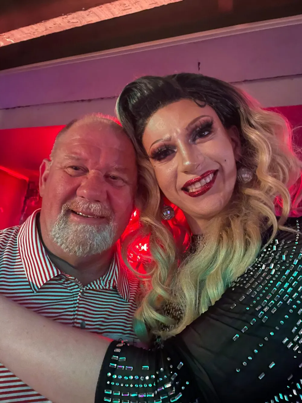 A man and a drag queen with glamorous makeup and a sequined outfit are posing together for a selfie with a red lit background