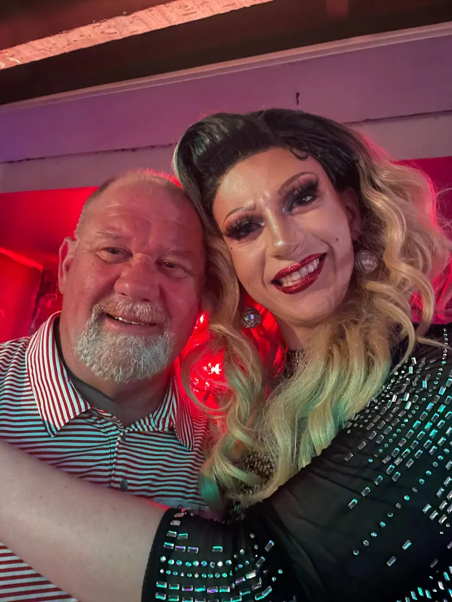 A man and a drag queen with glamorous makeup and a sequined outfit are posing together for a selfie with a red lit background.