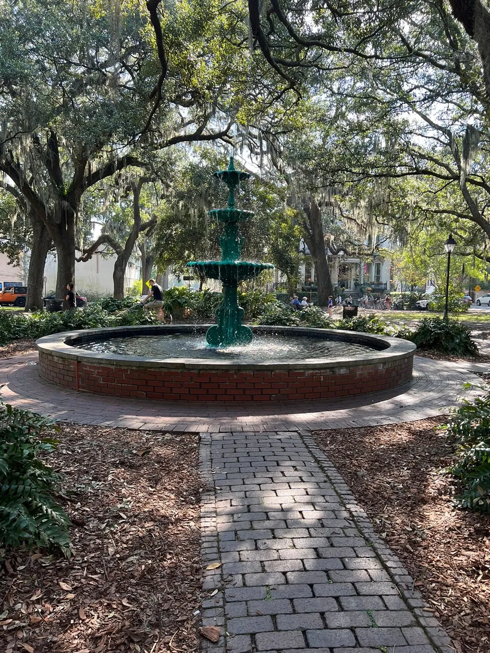 A serene public park scene featuring a multi-tiered fountain surrounded by lush trees draped with Spanish moss brick pathways and people enjoying the outdoors