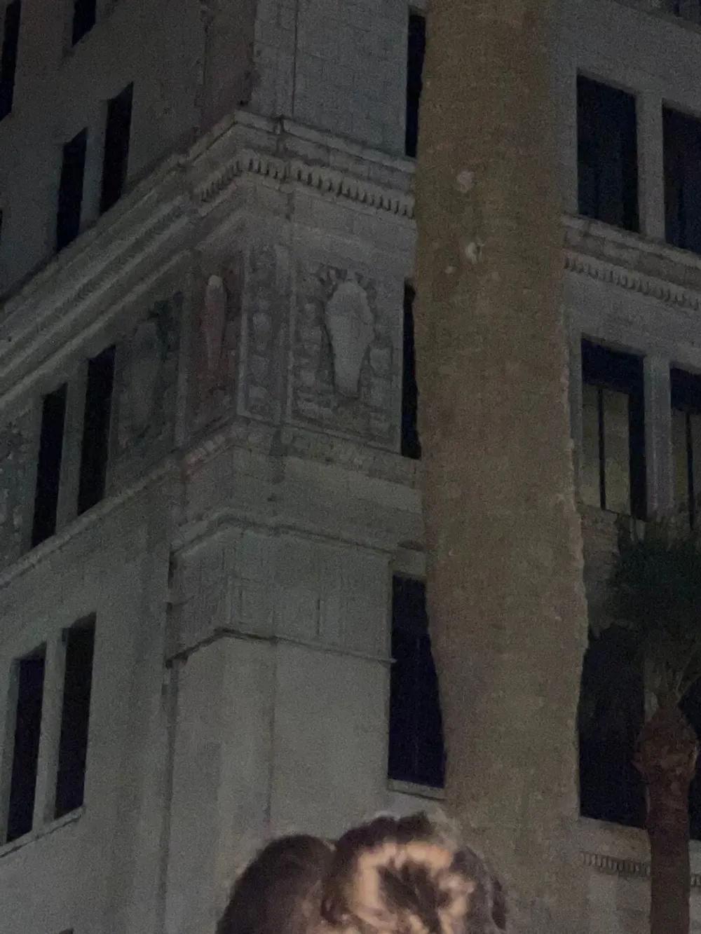 The image captures the corner of a building at night with architectural details and sculptural reliefs visible taken from a low angle where the silhouette of a persons head is seen in the bottom of the frame