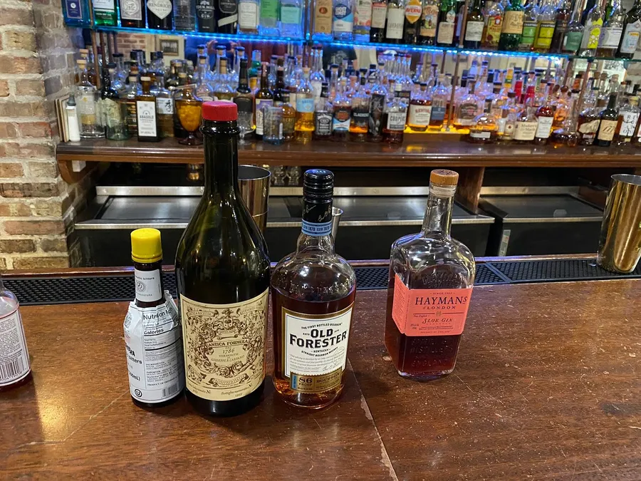 Three bottles of alcohol—vermouth, bourbon, and gin—are prominently displayed on a bar counter with an array of spirits in the background.