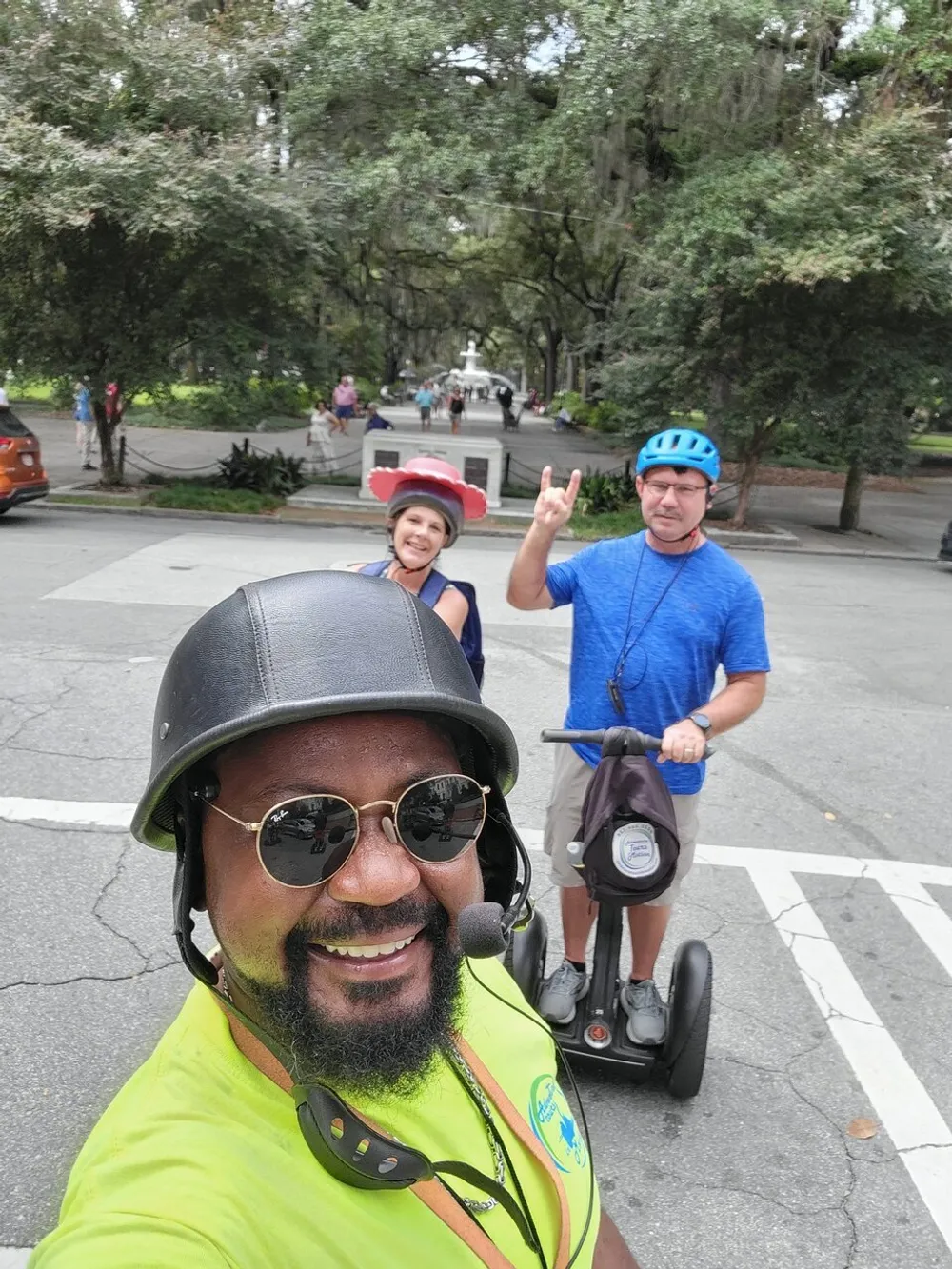 Three individuals are posing for a selfie on a sunny day with two of them standing behind on Segways and the person in the foreground wearing a helmet and sunglasses giving a thumbs-up sign