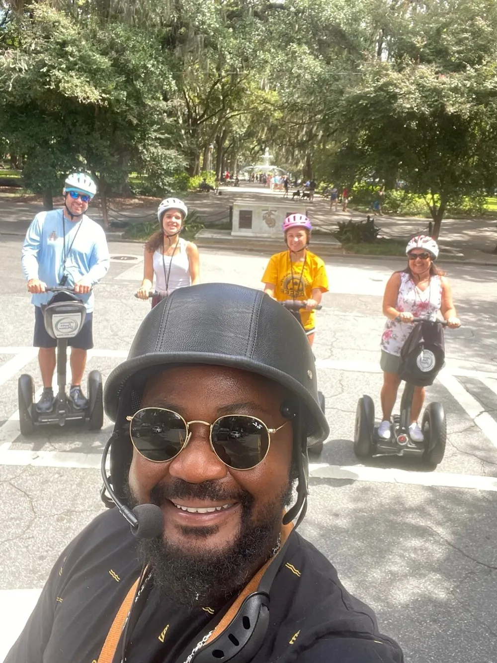 A group of people with one taking a selfie wearing helmets and riding Segways on a tree-lined street