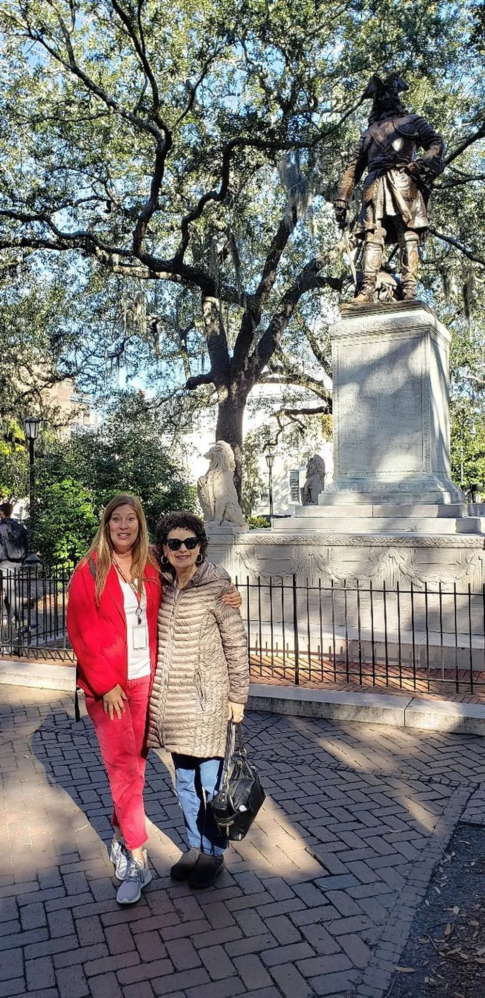 Two individuals are posing for a photo in a park with a statue on a pedestal and trees draped with Spanish moss in the background