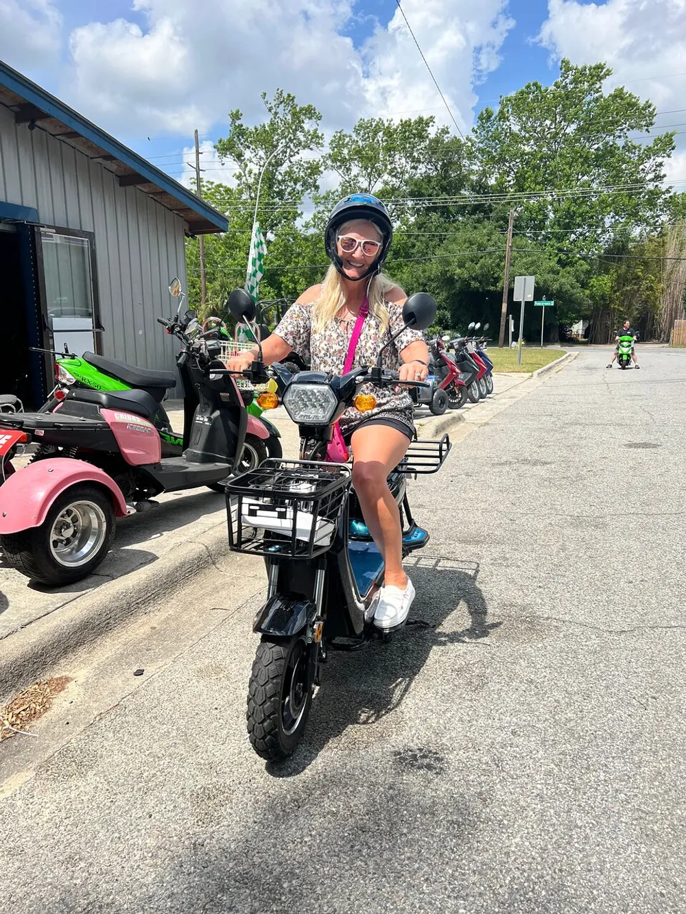 A person wearing a helmet and sunglasses is smiling while sitting on a black moped with a basket with several scooters in the background on a sunny day