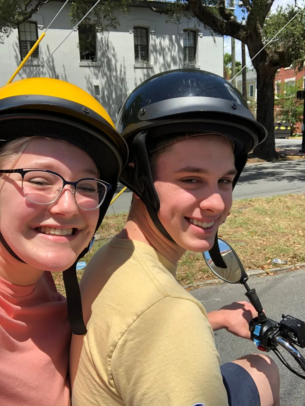 Two smiling people wearing helmets are taking a selfie likely on a sunny day possibly before or after a ride on a motorcycle or scooter