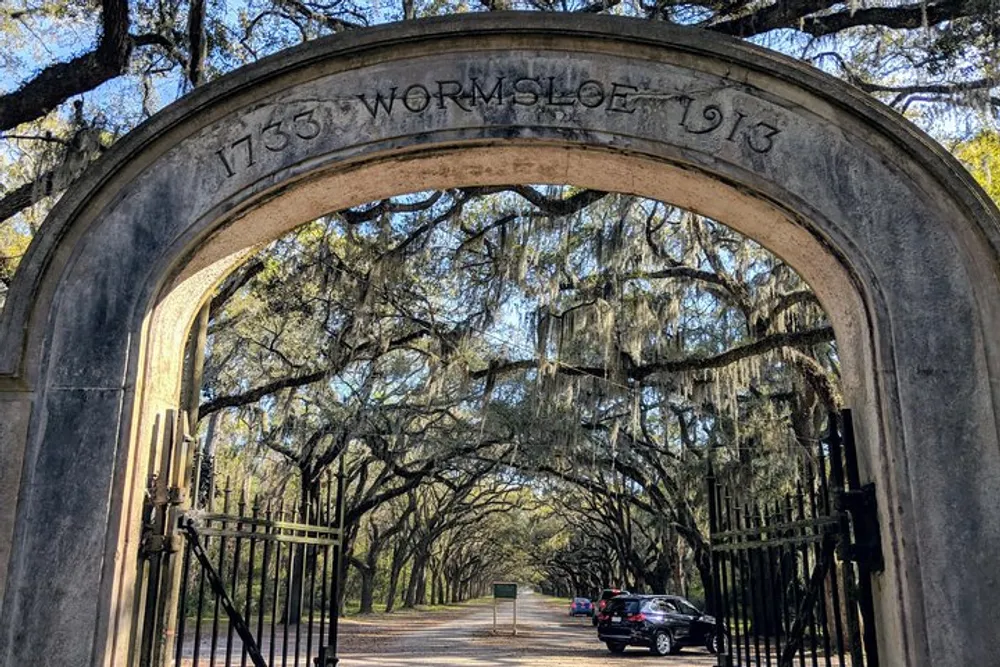 The image shows an ornate stone archway inscribed with 1733 WORMSLOE 1913 leading into a picturesque avenue lined with live oak trees draped with Spanish moss with a car driving through and another parked nearby
