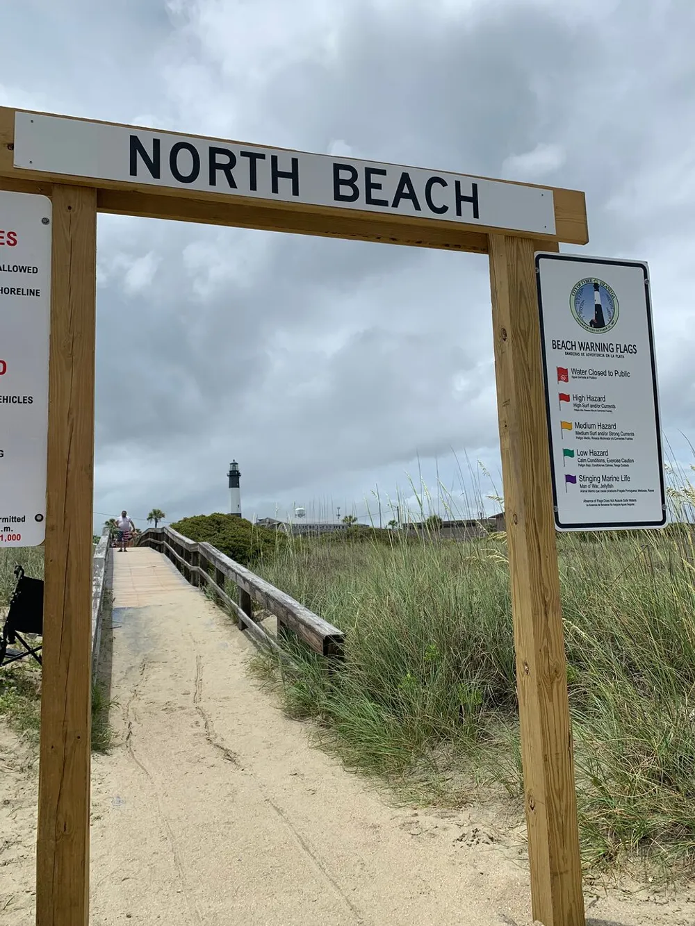 The image shows the entrance to North Beach with a signpost and a beach warning flags board a sandy pathway leading to the beach and a lighthouse in the background under an overcast sky