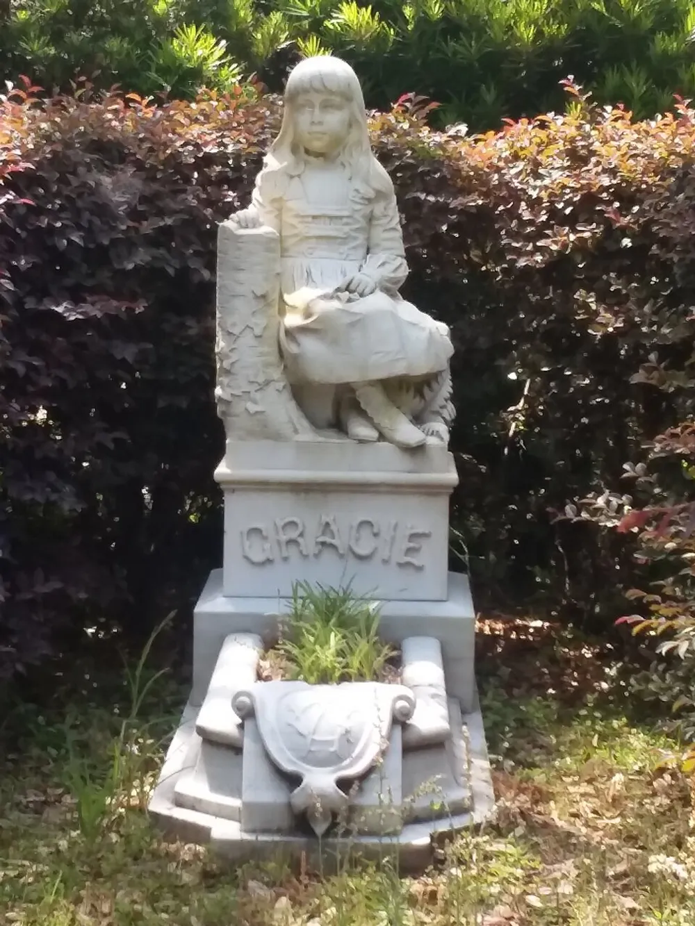 The image shows a stone statue of a young girl named Gracie sitting next to a pedestal with a serene expression nestled among greenery