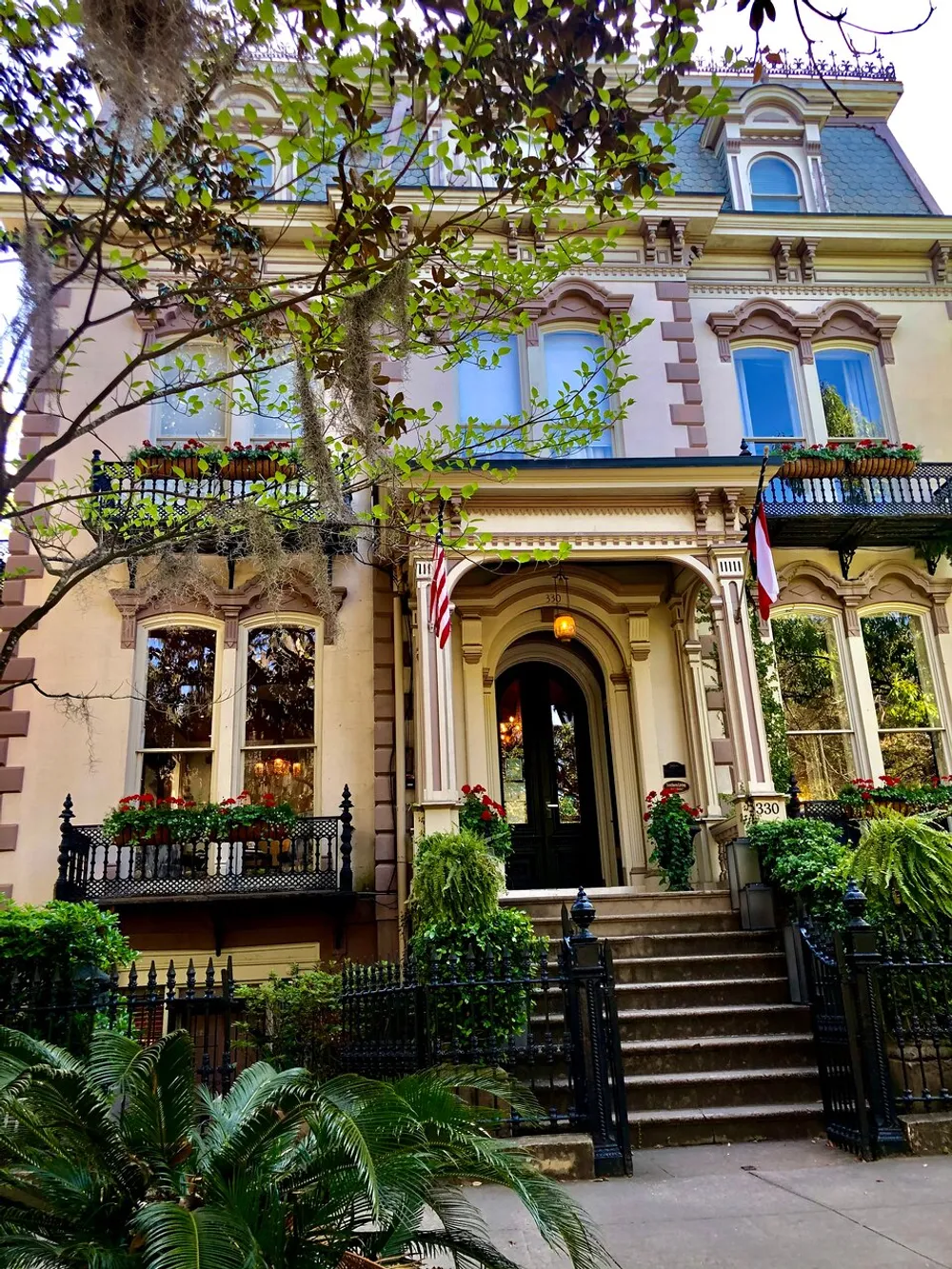 The image shows a charming multistory Victorian house with intricate ironwork on the balconies vibrant flowerboxes and an American flag nestled among greenery and mature trees
