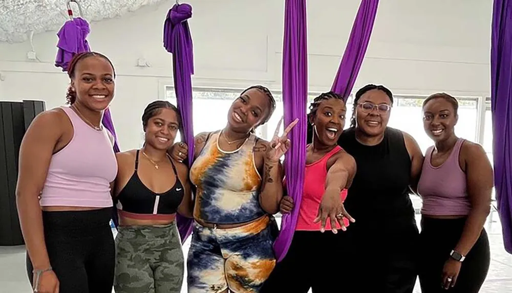 A group of six smiling women in workout attire poses for a photo in a studio with purple aerial silks hanging in the background